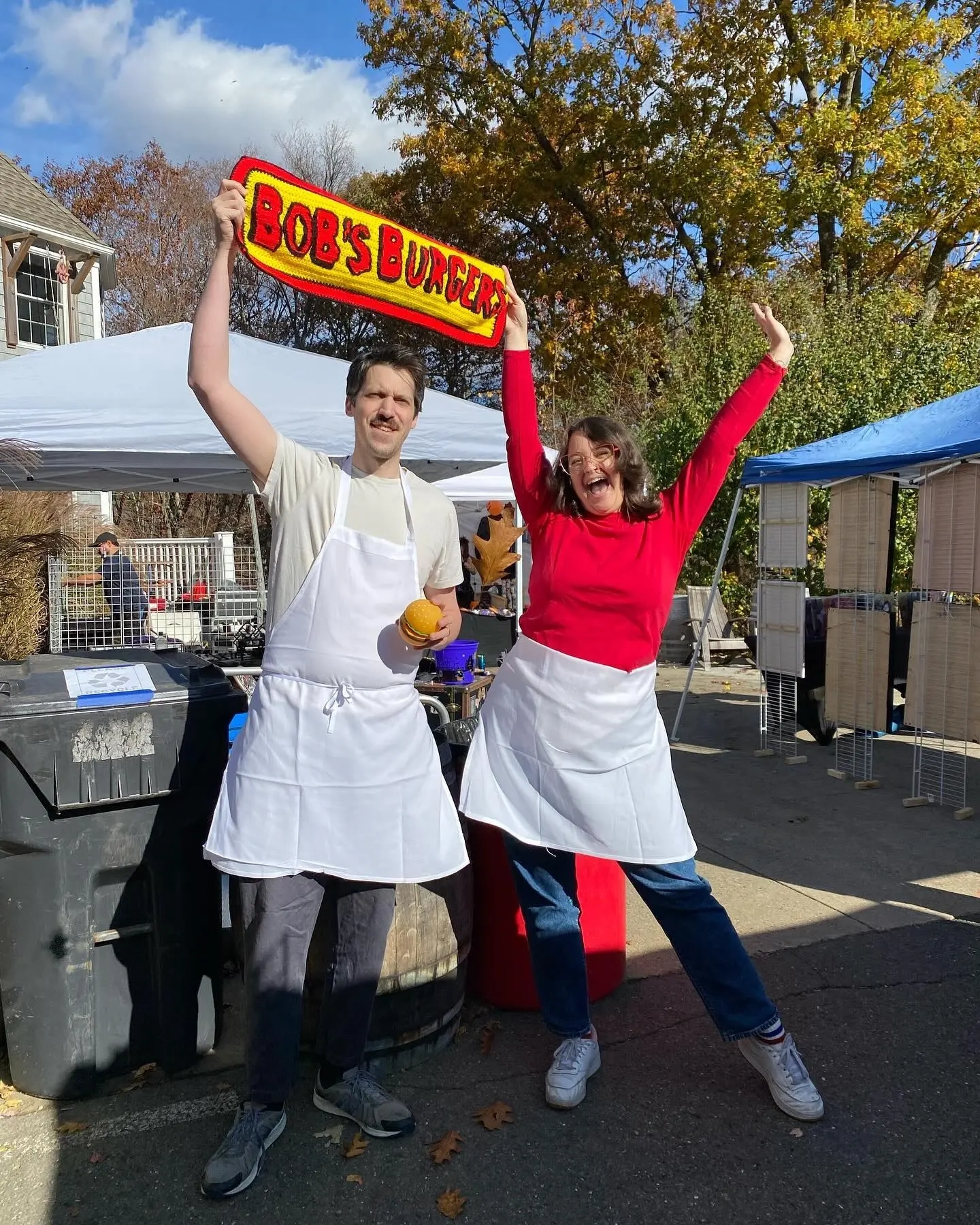 Couple in white aprons and casual clothes holding Bob's Burgers sign as restaurant workers costume 3