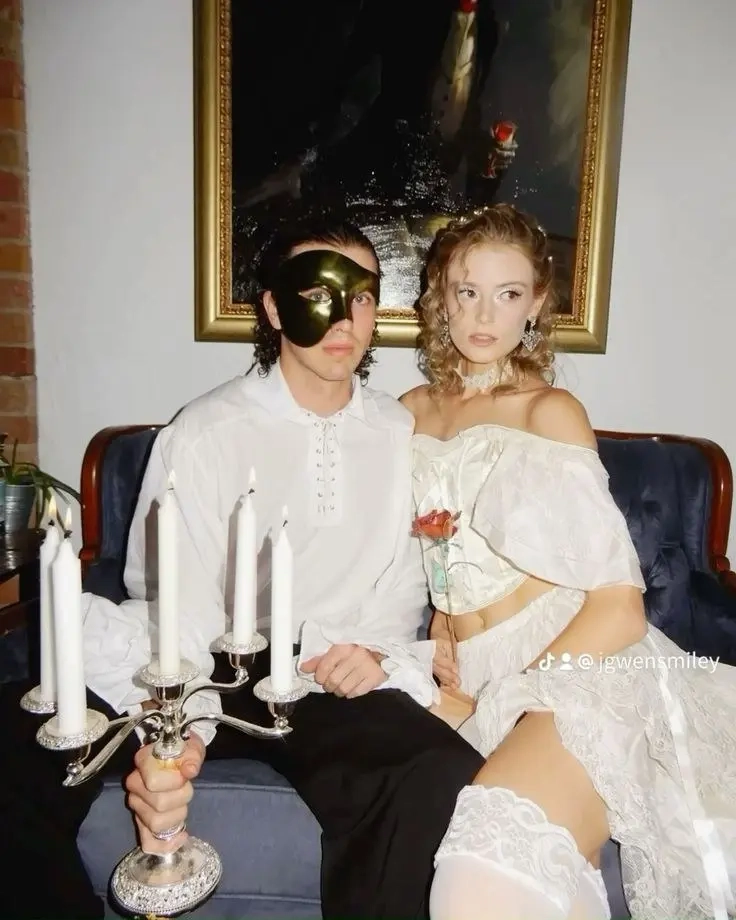 man in white shirt with gold mask holding candle next to woman in white off-shoulder dress with pearls outfit 3