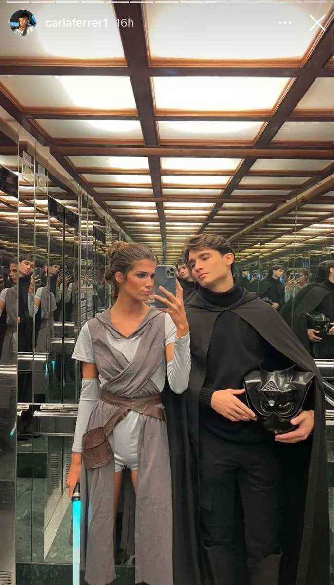 woman in beige wrapped dress with belt next to man in black hooded outfit taking elevator mirror selfie outfit 9