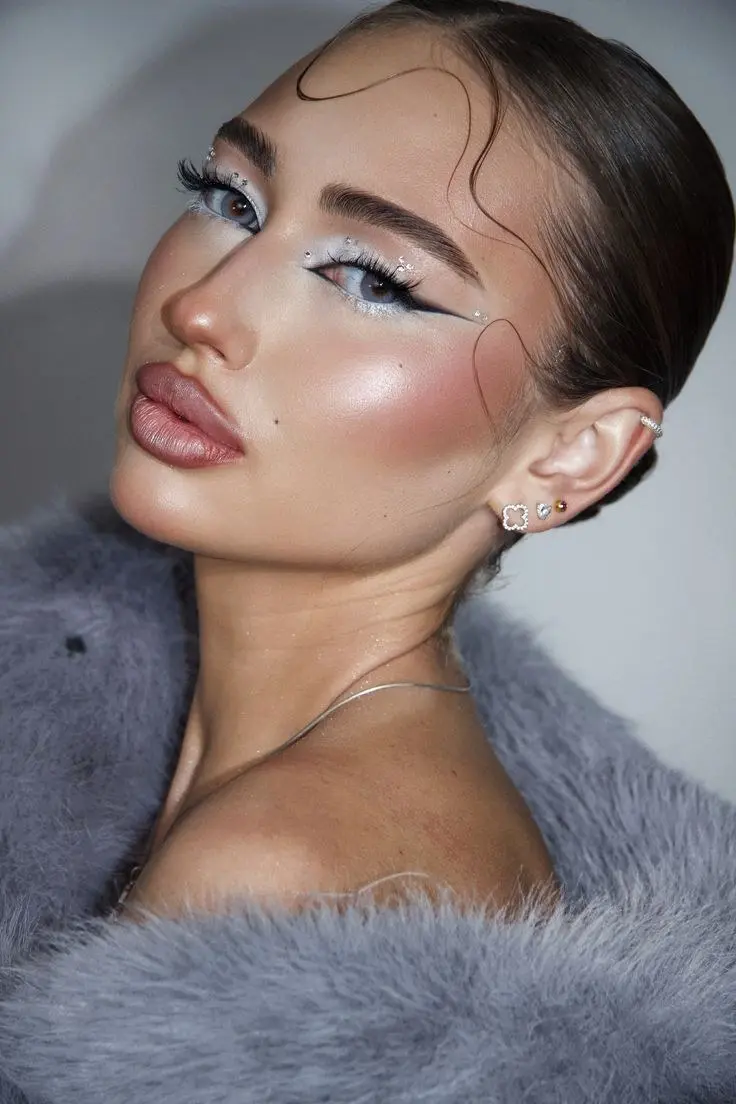 brunette woman with white ice blue eyeshadow graphic liner wing crystal gems scattered near eye heavy warm blush glazed fur collar cold girl makeup look 10