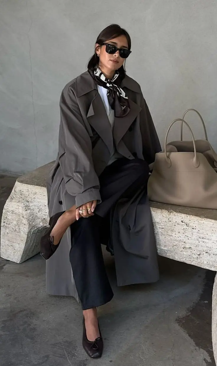 woman in grey trench coat black wide leg trousers light blue shirt printed neck scarf dark loafers beige tote bag seated on stone bench fall work outfit 1