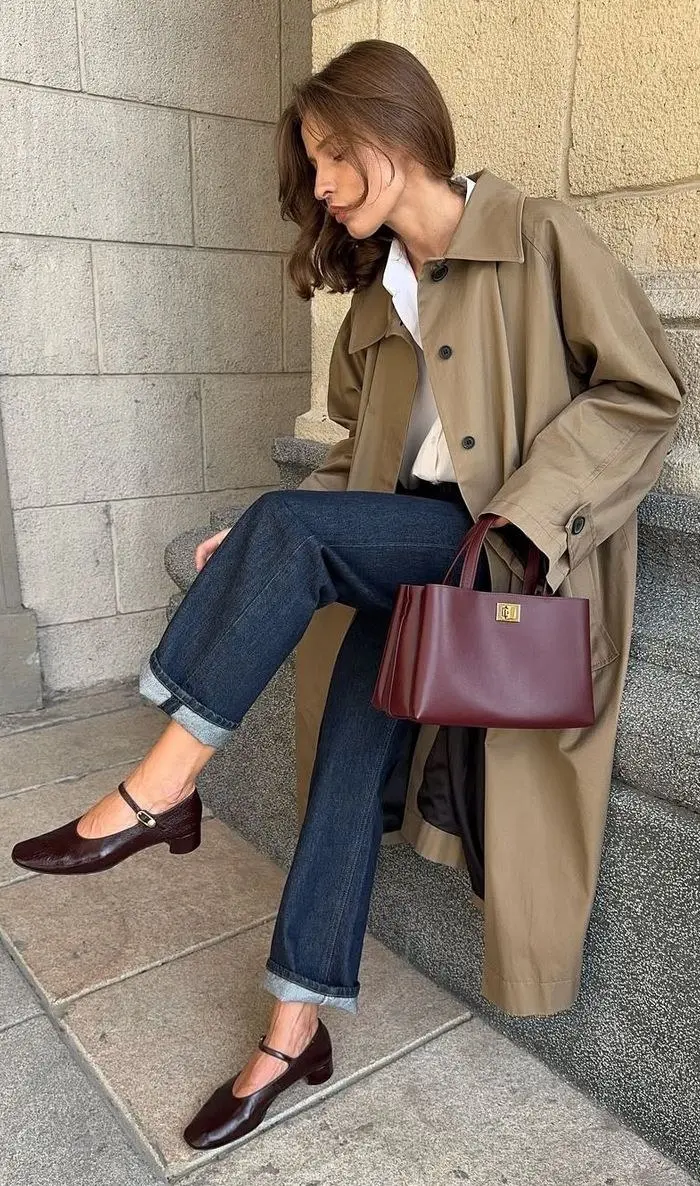 woman in camel trench coat white shirt dark indigo cuffed straight jeans burgundy croc Mary Jane low heels burgundy structured tote bag seated on stone steps fall work outfit 14