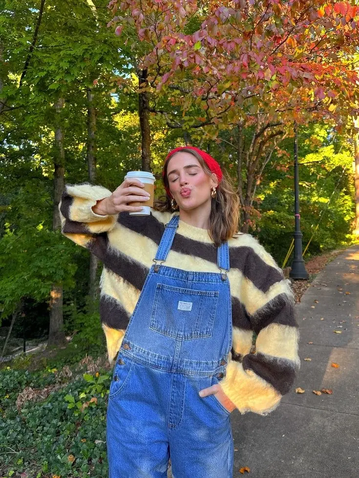 woman outdoors in yellow brown striped mohair sweater under light wash denim overalls red headband gold earrings autumn trees pumpkin patch outfit 18