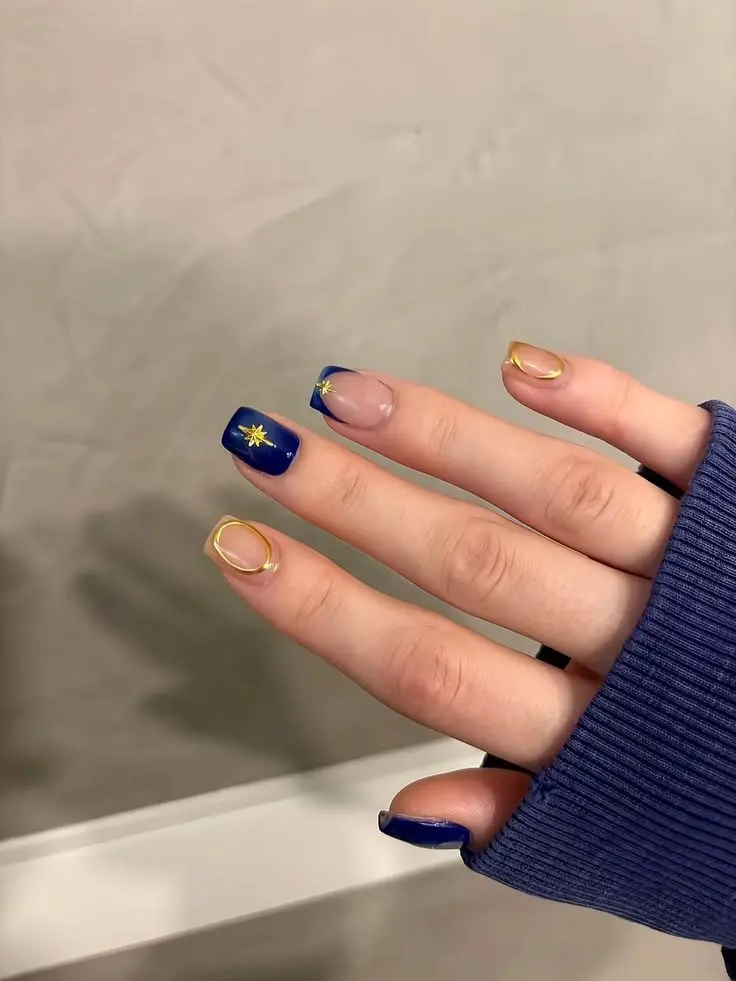 short square nails in deep cobalt navy with gold French tips on two fingers and gold starburst art on one accent nail, hand in blue sleeve, photo 6