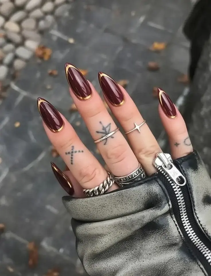 long almond nails in deep wine burgundy gel with gold chrome outline along sides and tip, high gloss finish, silver rings on tattooed hands, photo 8