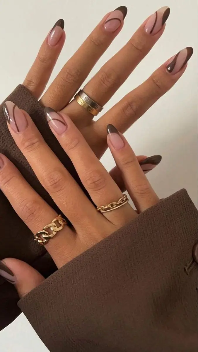 medium almond nails with sheer nude blush gel base and dark chocolate brown flowing swirl line art brushstrokes, one nail with rhinestone accent, gold chain rings, photo 19