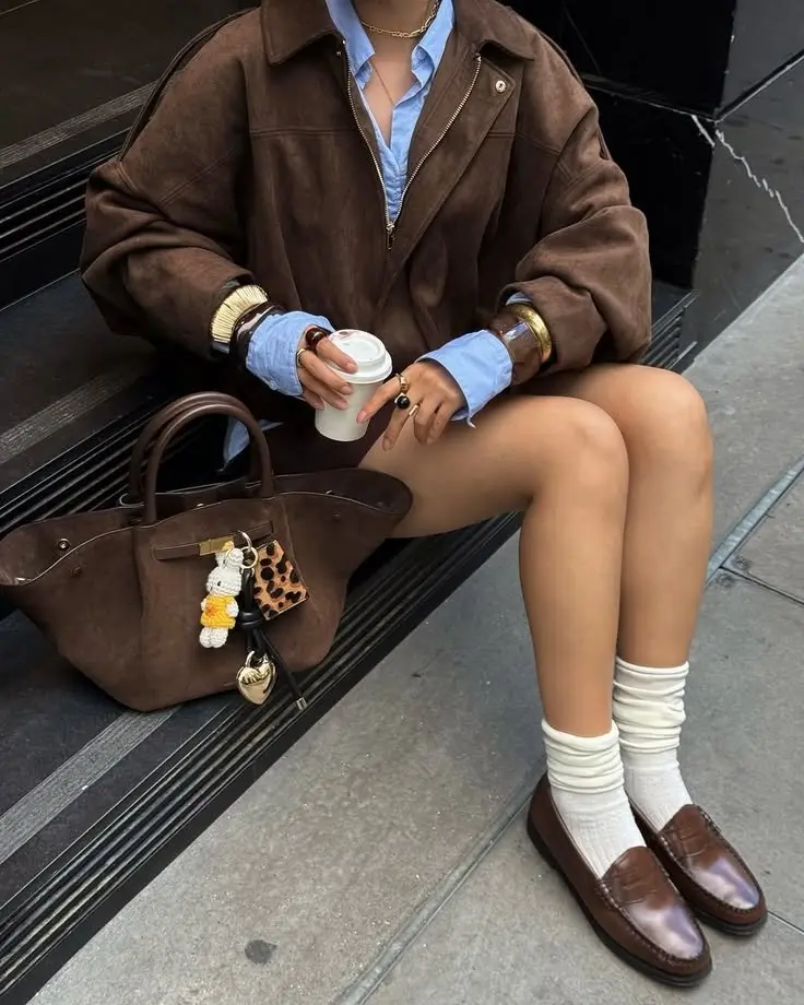 woman sitting on steps in chocolate brown oversized suede zip jacket light blue oxford shirt white crew socks brown penny loafers gold cuff bracelets and brown structured tote bag outfit 14