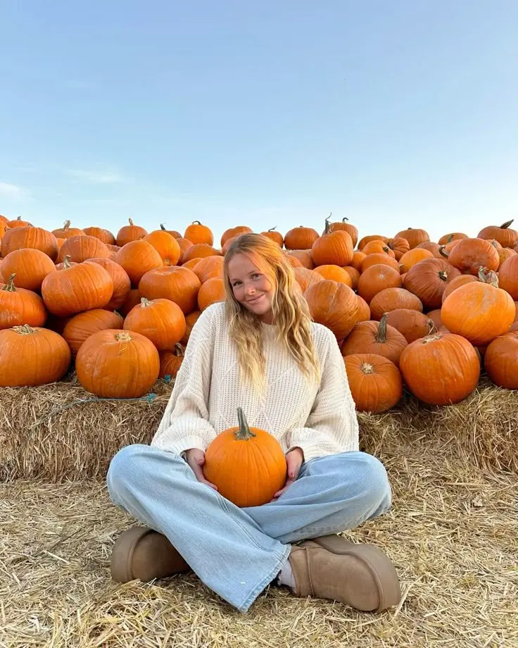 woman in cream oversized cable knit sweater light wash wide leg jeans taupe UGG platform boots sitting on hay bales holding pumpkin at pumpkin farm outfit 16