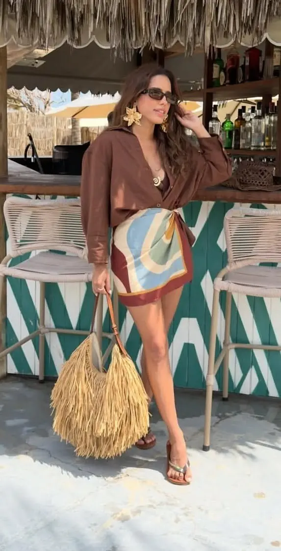 woman in brown oversized linen shirt open over bikini top with colorful printed sarong wrap skirt raffia fringe tote bag and flat sandals at beach bar outfit 2