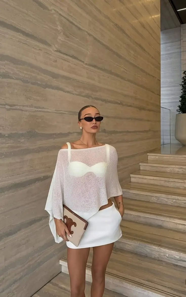 woman in sheer white open knit oversized top over white bandeau and white mini skirt holding neutral structured clutch and wearing small oval sunglasses in hotel lobby outfit 3