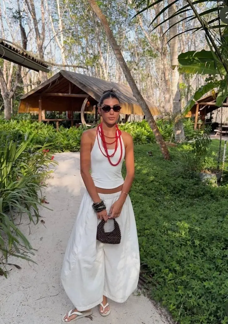 woman in white halter crop top and white wide leg linen pants with red beaded layered necklaces small dark woven bag white flip flops in tropical garden setting outfit 5