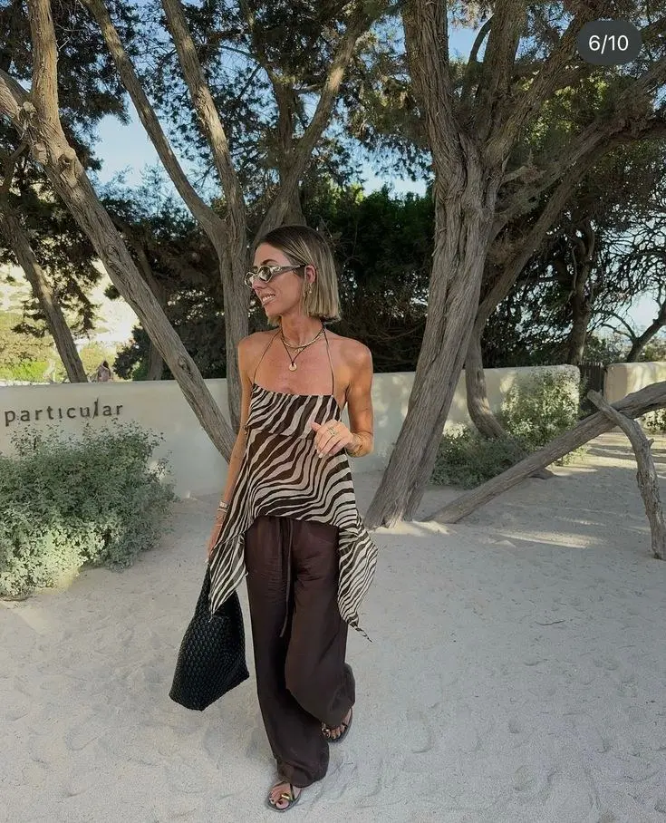 woman in zebra print asymmetric halter top and dark brown wide leg linen trousers with large black woven tote bag and flat strappy sandals on sandy path outdoor setting outfit 6