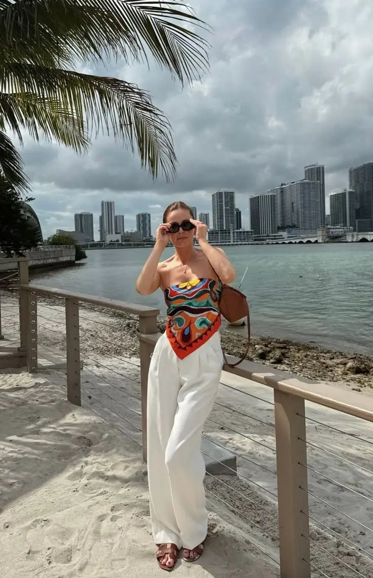 woman in colorful printed strapless bandeau top and white wide leg trousers with tan structured shoulder bag and flat sandals by waterfront with city skyline background outfit 7