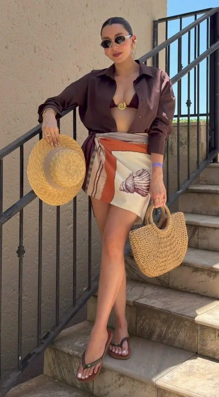 woman in dark brown oversized shirt open over dark bikini top with printed shell motif sarong wrap skirt round woven straw bag and flat straw hat on outdoor stone steps outfit 9