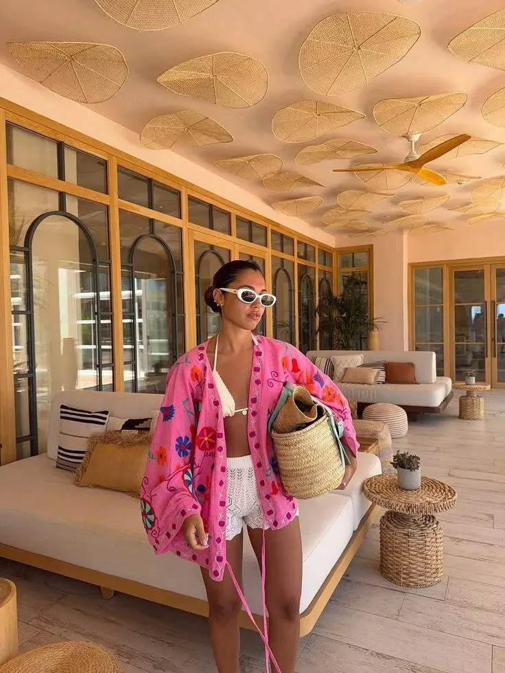 woman in pink large floral embroidered kimono coverup over white bikini top and white crochet shorts holding woven market basket bag in terrace resort interior outfit 10
