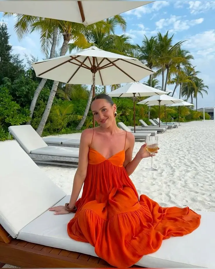 woman in tiered rust orange gauze maxi dress with spaghetti straps sitting on white beach lounger with palm trees and white umbrellas in background outfit 11