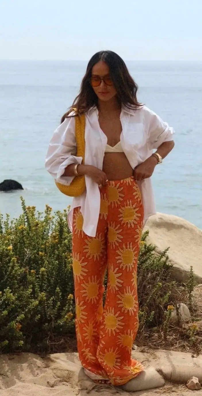 woman in orange and gold sun print wide leg pants white bikini bra top and open white oversized linen shirt with woven straw shoulder bag standing on coastal cliffside outfit 14