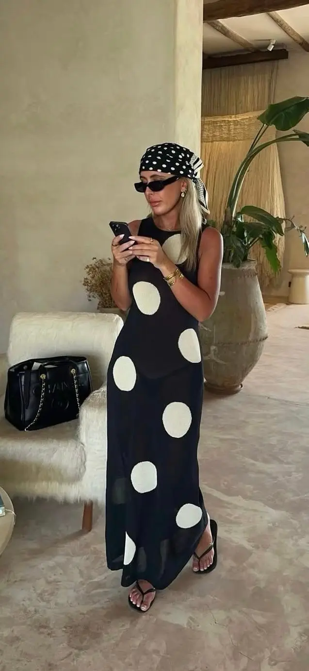 woman in black and white large polka dot sleeveless semi-sheer knit maxi dress with polka dot hair scarf wrap black chain shoulder bag black flip flops and gold jewelry in warm interior outfit 16