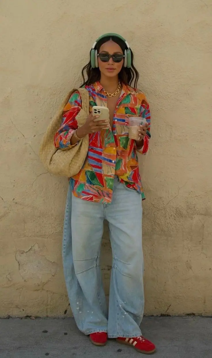 woman in colorful abstract print button-down shirt light wash wide leg jeans red adidas sneakers woven bag green headphones sunglasses outfit 1