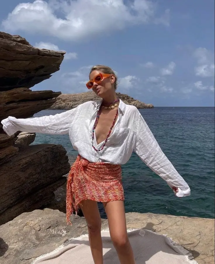 woman in oversized white linen shirt printed orange and red wrap mini skirt layered beaded necklaces orange sunglasses standing on coastal rocks ocean behind outfit 7