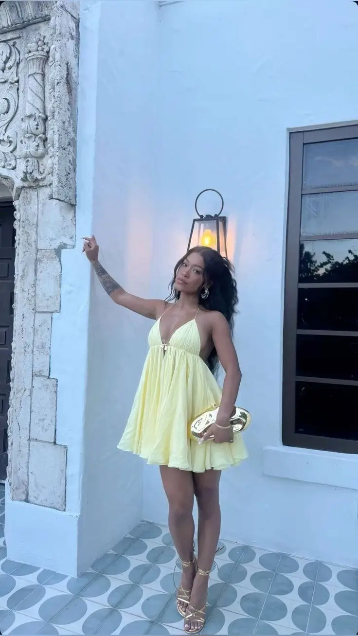 woman in yellow spaghetti strap halter babydoll mini dress gold oval metallic clutch gold strappy heeled sandals standing outside white stucco building at dusk outfit 8
