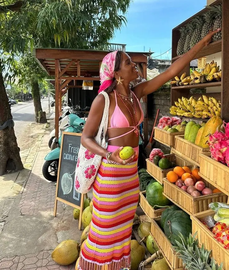 woman in pink bikini top multicolor striped crochet maxi skirt crochet tote bag printed pink headscarf gold layered necklaces reaching for fruit at outdoor market outfit 9