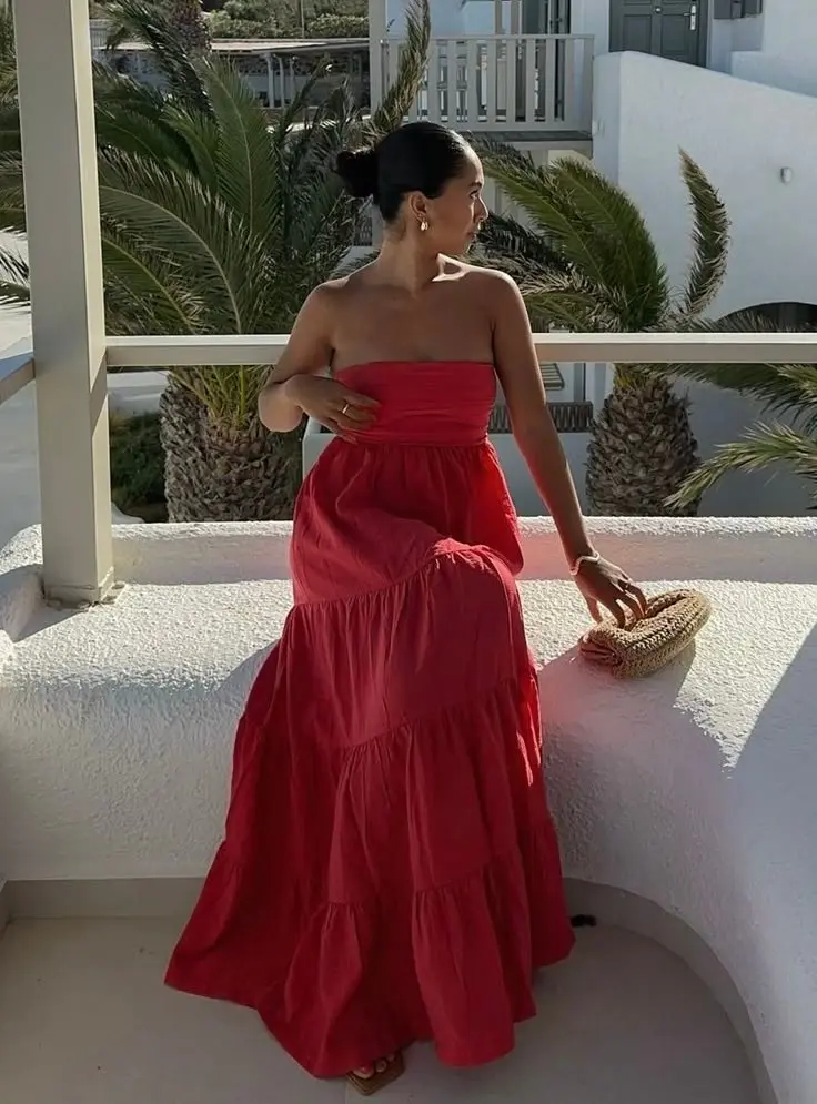 woman in red strapless tiered maxi dress small woven circular clutch gold earrings standing on white balcony with palm trees and ocean view outfit 12