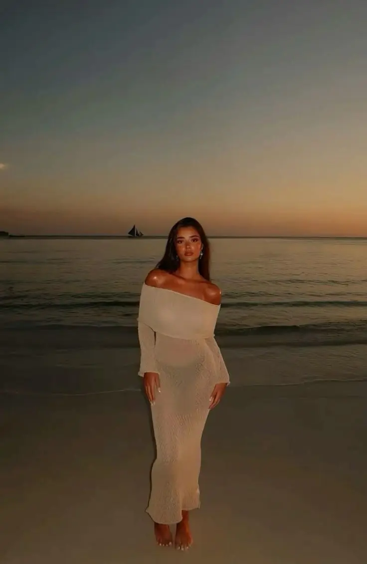 woman in cream off-shoulder long sleeve crochet texture maxi dress barefoot on beach at sunset ocean and sailboat behind dramatic sky outfit 17