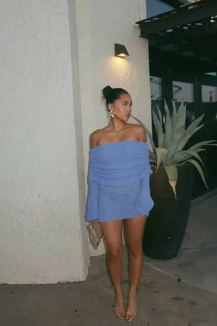 woman in periwinkle blue off-shoulder ruched mini dress crystal statement earrings high bun gold metallic clutch clear strappy heeled sandals standing outside venue at dusk outfit 18