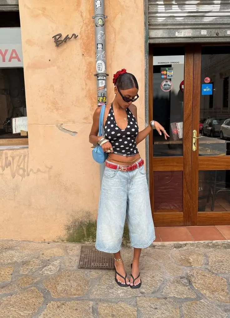 woman in black and white polka dot halter crop top wide leg light wash denim jorts red embellished belt blue small shoulder bag flip flops silver bangles red hair scrunchie on cobblestone street outfit 19