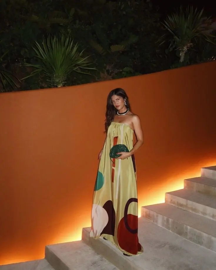 woman in yellow abstract geometric print strapless full-length maxi dress green woven clutch layered pearl and chain necklaces standing on lit steps at night tropical garden resort outfit 20