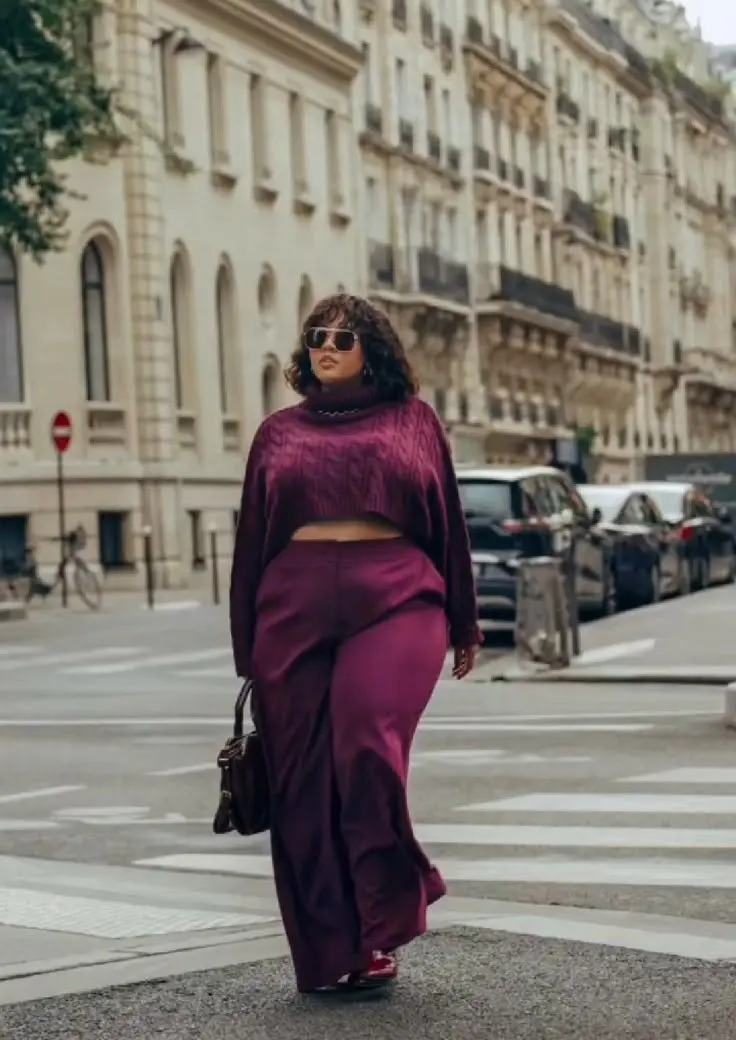 Curvy fashion blogger in deep burgundy tonal outfit — structured knit top, high-waist wide-leg pants, wool coat, fall foliage background