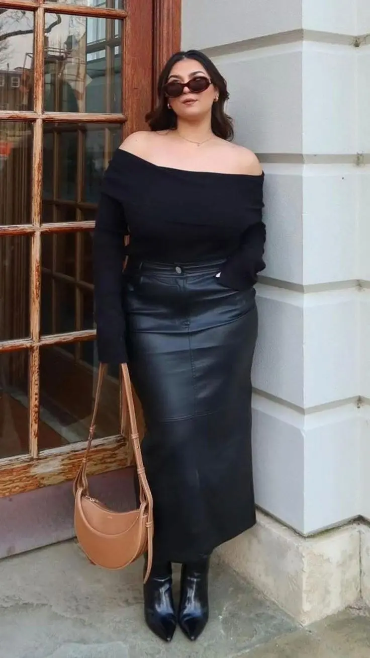 Curvy woman in black faux leather A-line midi skirt, cream ribbed turtleneck, ankle boots, city street fall setting