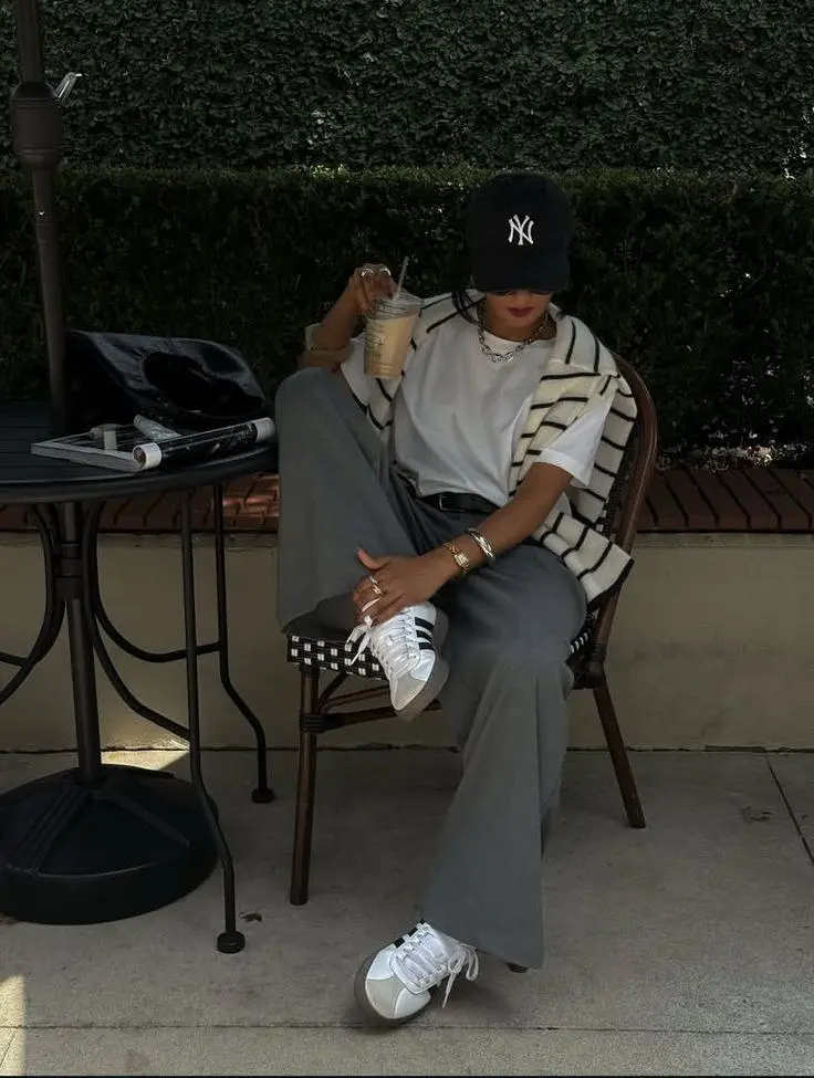 woman seated at outdoor cafe table wearing black NY cap white tee striped knit vest grey wide-leg trousers white Adidas Superstar sneakers gold layered necklace bracelet iced coffee outfit 2