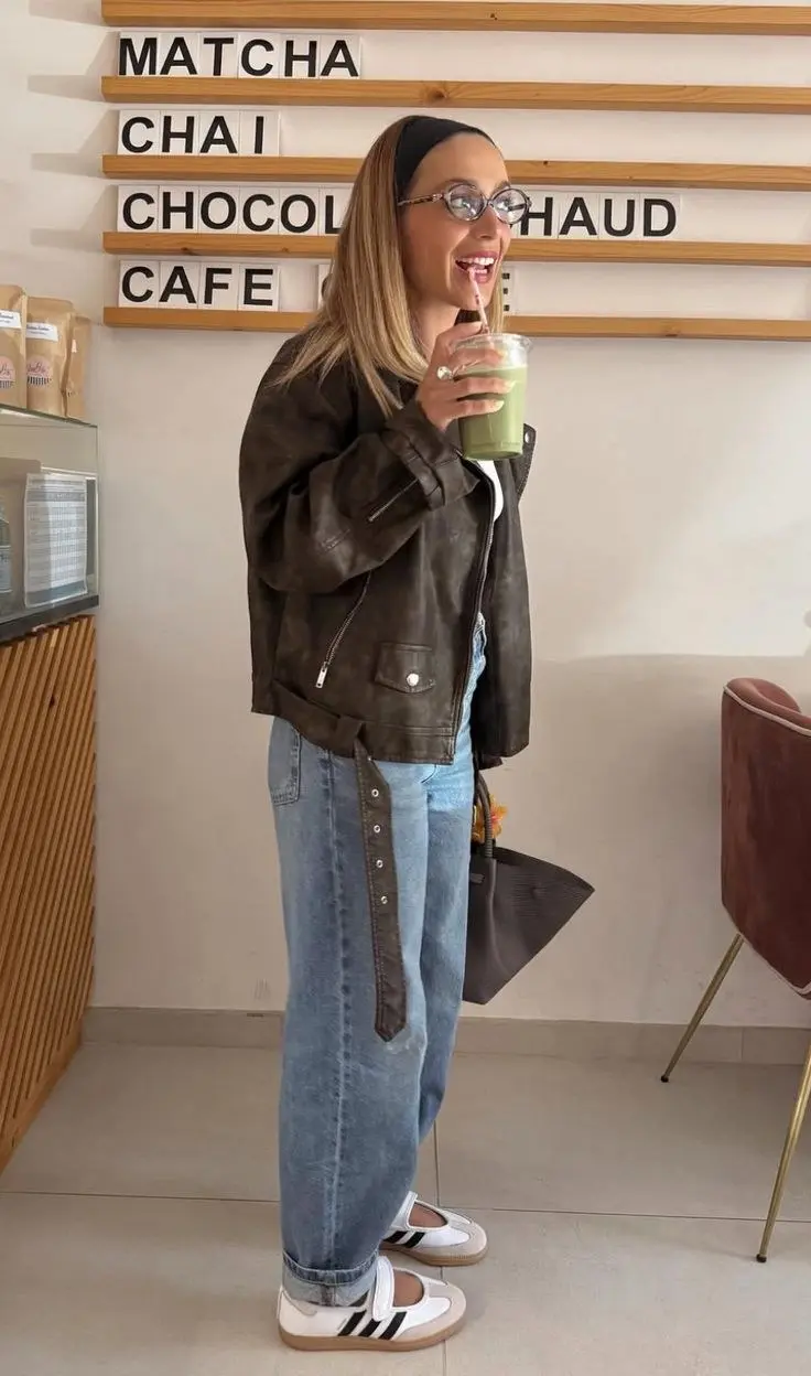 woman in brown distressed leather jacket baggy light-wash jeans white Adidas Samba sneakers black headband round glasses carrying canvas tote bag at cafe outfit 7