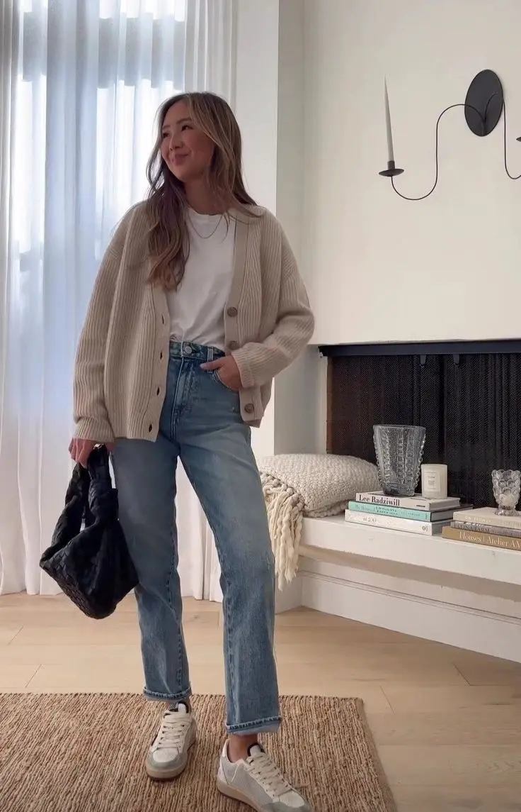 woman wearing beige oversized ribbed button cardigan white tee straight leg light-wash jeans white low-top sneakers black puffer bag indoor home setting outfit 8
