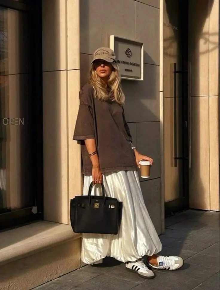 woman wearing oversized dark brown washed tee white pleated satin maxi skirt white Adidas sneakers beige cap large black structured tote bag street outfit 9