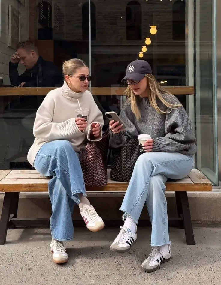 two women seated on bench wearing chunky turtleneck sweaters wide-leg light-wash jeans Adidas Samba white sneakers one in cream turtleneck one in grey turtleneck with NY cap and glasses coffee cups street outfit 11