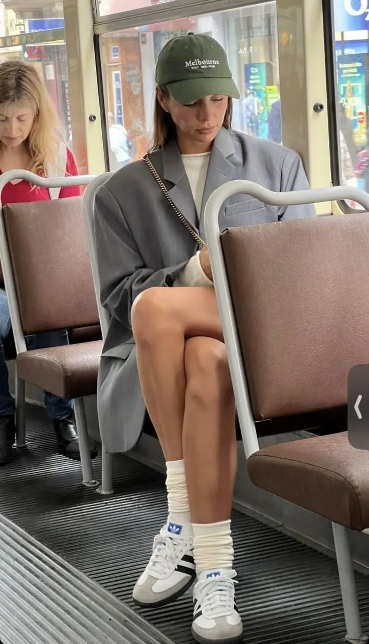 woman on bus wearing green Melbourne Golf Club cap oversized grey blazer as coat white mini dress white Adidas Samba sneakers white scrunched socks gold chain shoulder bag outfit 12
