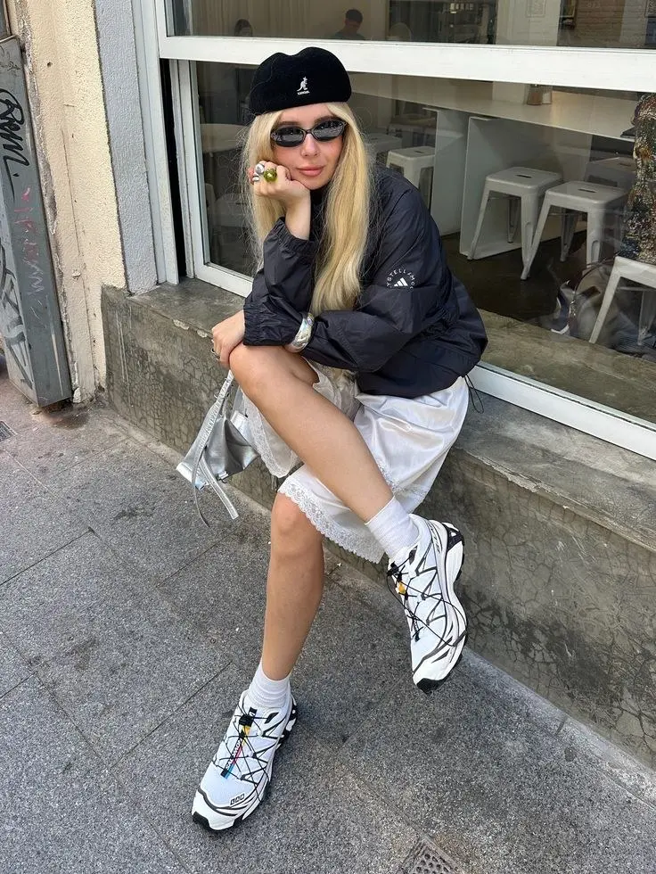 young woman sitting on street ledge wearing black velvet Kangol beret slim sunglasses black sport windbreaker white lace-trim slip shorts white chunky trail sneakers silver metallic mini bag rings outfit 15