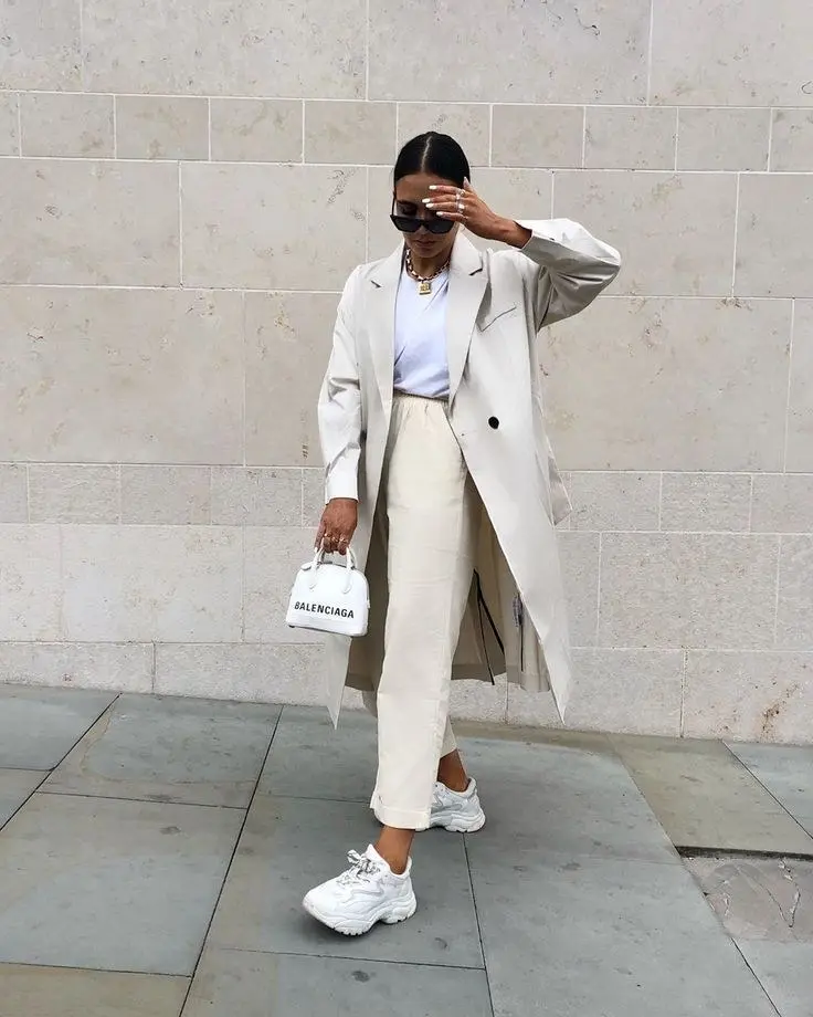 woman in cream longline trench coat white wide-leg trousers light blue fitted tee white chunky sneakers small white Balenciaga mini bag gold chain necklace sunglasses outdoor street outfit 16
