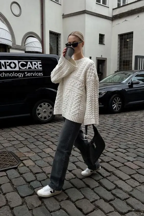 woman in oversized cream cable knit sweater dark grey straight jeans black shoulder bag and white Veja sneakers walking on cobblestone outfit 8