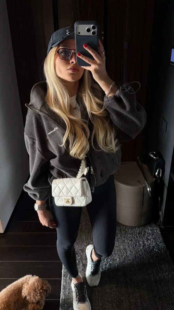 blonde woman in oversized dark charcoal zip hoodie black leggings white quilted crossbody bag black cap square sunglasses On Cloud sneakers mirror selfie outfit 2