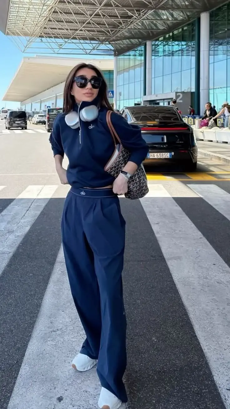 brunette woman in matching navy half-zip sweatshirt and wide leg jogger pants white On Cloud sneakers brown tote bag sunglasses standing outside airport terminal outfit 3