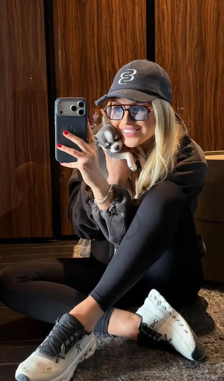 blonde woman sitting on floor in all-black zip hoodie and leggings On Cloud sneakers black baseball cap large square glasses holding puppy mirror selfie outfit 7