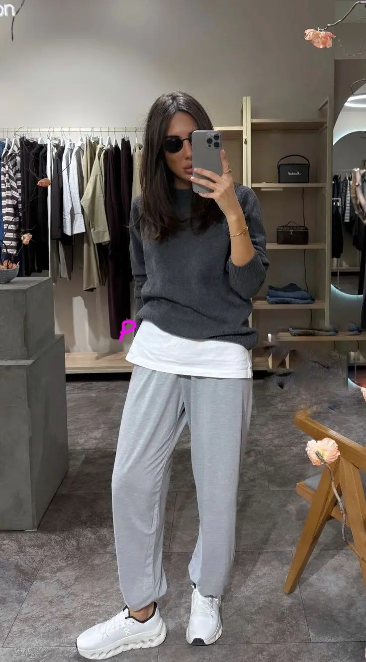 brunette woman in store mirror wearing charcoal crewneck sweater over white tee light grey relaxed jogger trousers white and black On Cloud sneakers sunglasses gold bracelet outfit 12