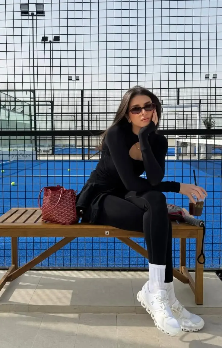 brunette woman in all-black fitted long sleeve turtleneck athletic top and leggings white On Cloud sneakers white crew socks small dark sunglasses sitting on bench near blue padel court red bucket bag outfit 14