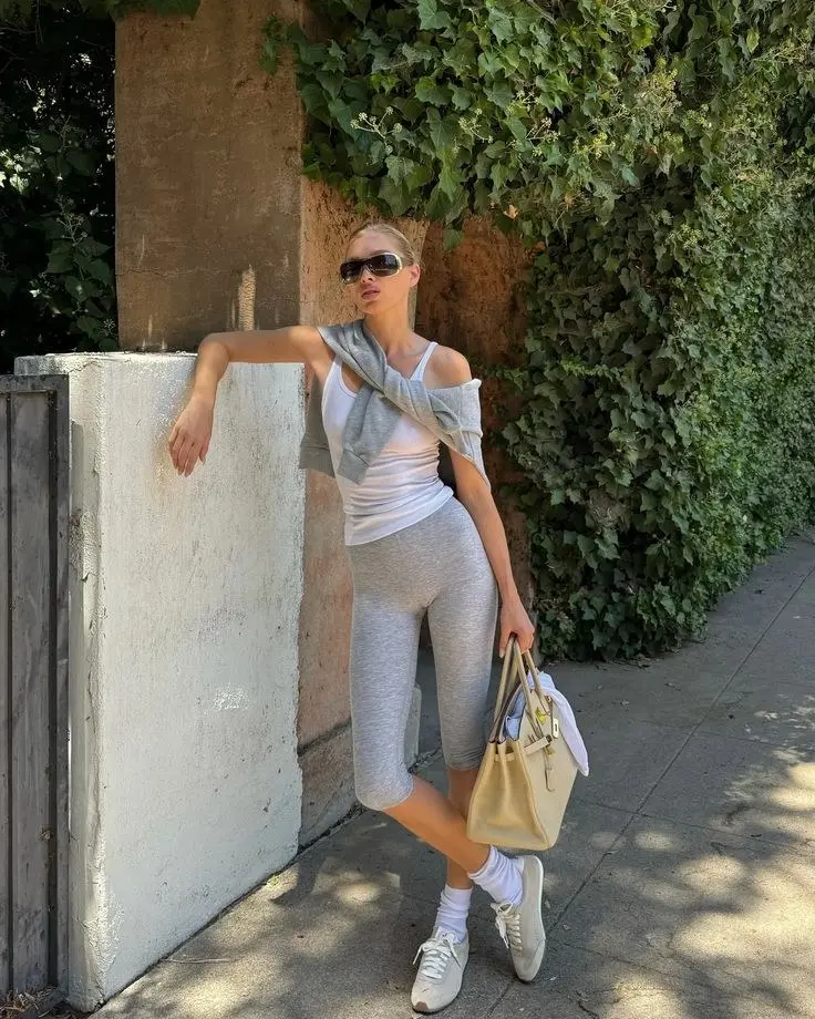 woman leaning against wall in white tank top grey bike shorts sweatshirt draped off shoulder cream structured tote cream Alo sneakers white socks shield sunglasses outfit 5