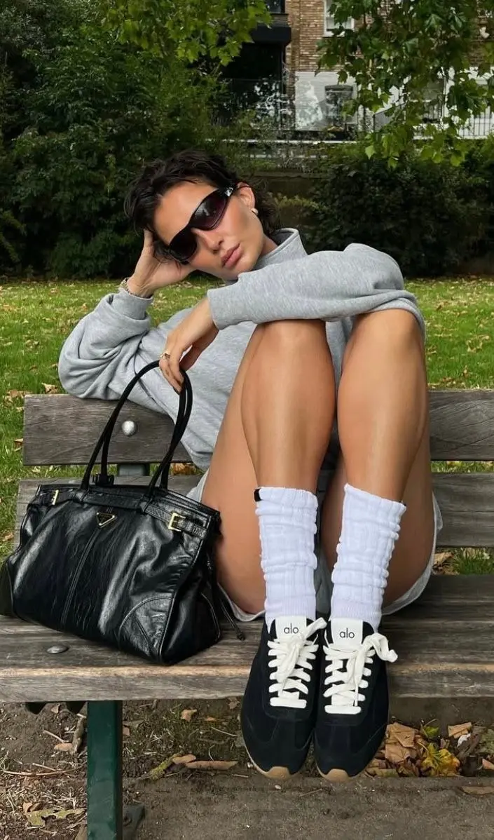 woman sitting on park bench in grey oversized hoodie bare legs white slouchy socks black Alo sneakers black patent structured bag sunglasses outfit 8