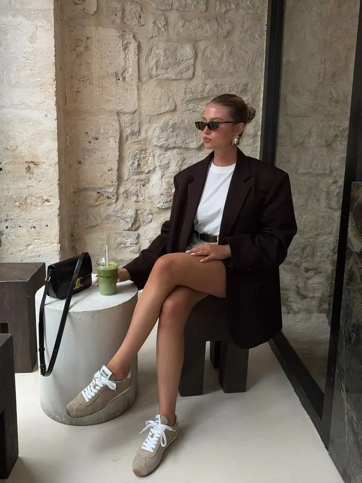 woman seated at cafe table in dark chocolate oversized blazer white tee black shorts tan suede Alo sneakers small black crossbody bag pearl earrings hair bun sunglasses outfit 9